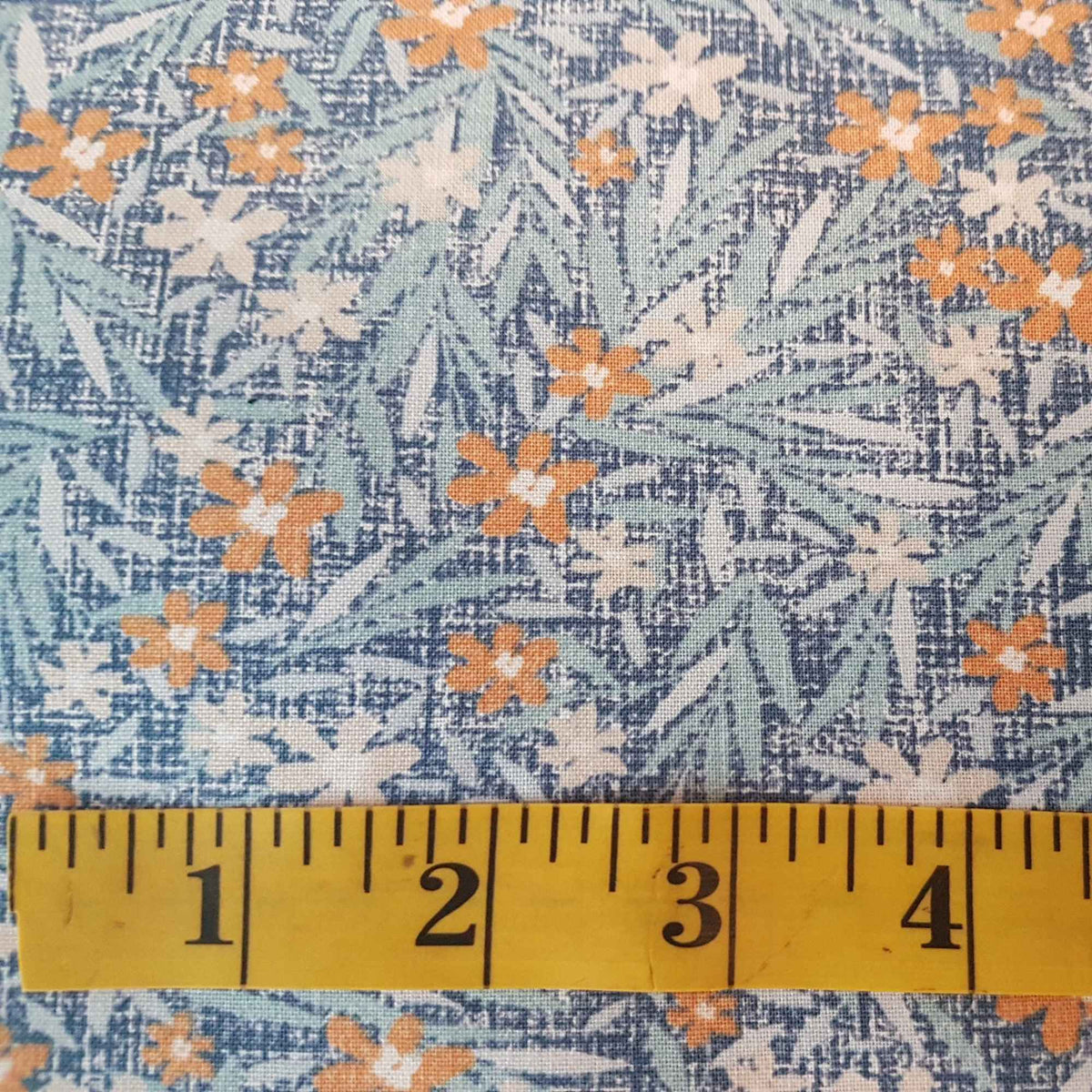 Cotton fabric with small orange flowers, cream accents, and green leaves on a blue textured background with ruler for scale.