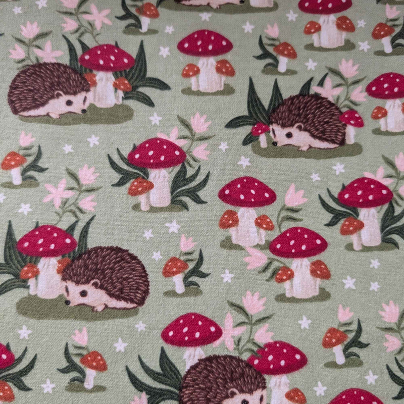 Cotton flannel fabric with woodland hedgehogs, red mushrooms, and greenery on a green background. Brushed soft, OEKO-TEX certified, 44 inches wide.