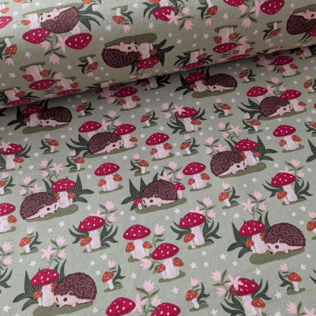 Cotton flannel fabric with woodland hedgehogs, red mushrooms, and greenery on a green background. Brushed soft, OEKO-TEX certified, 44 inches wide.