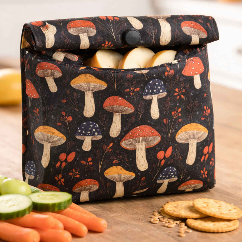 Reusable snap-closure snack bag made with red and golden mushroom print waterproof PUL fabric on black background, filled with apple slices and shown with crackers and vegetables on a wooden table.