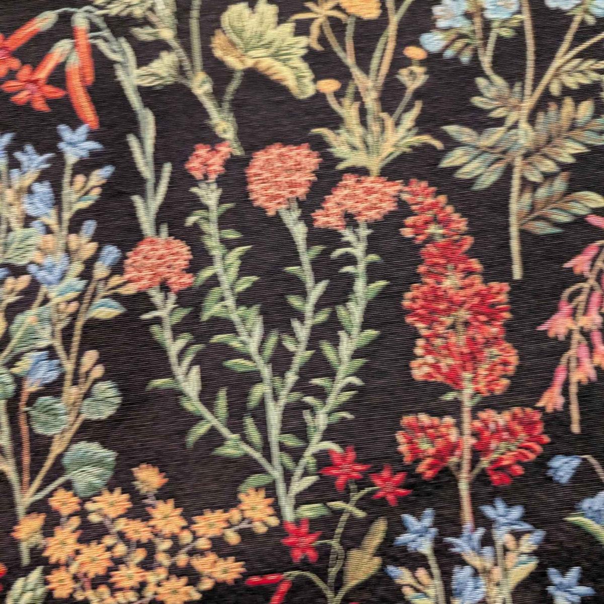 Botanical wildflower Gobelin tapestry fabric with red, coral, blue and yellow flowers and sage green leafy stems on a black background.