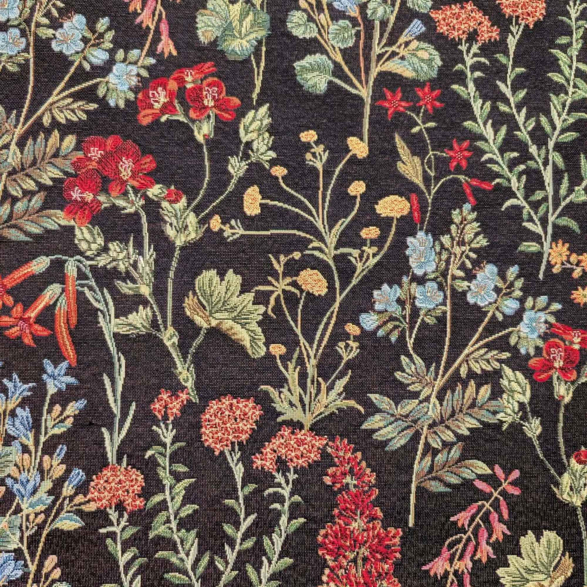 Botanical wildflower Gobelin tapestry fabric with red, coral, blue and yellow flowers and sage green leafy stems on a black background.