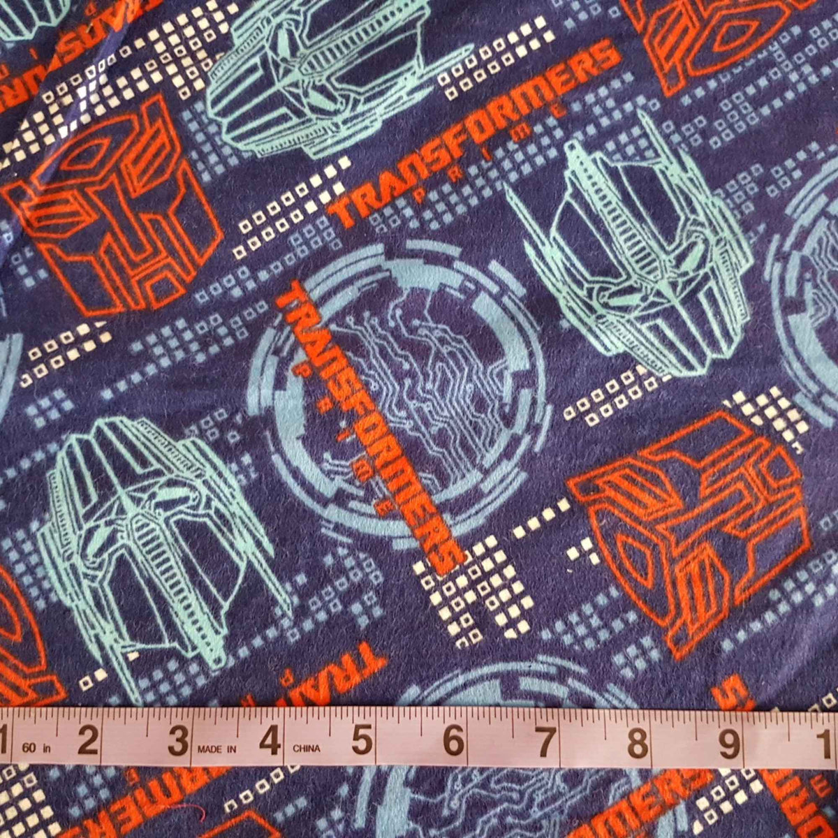 Transformers cotton flannel fabric with colorful robot designs and logos on a dark background, 44-inch wide kids character fabric