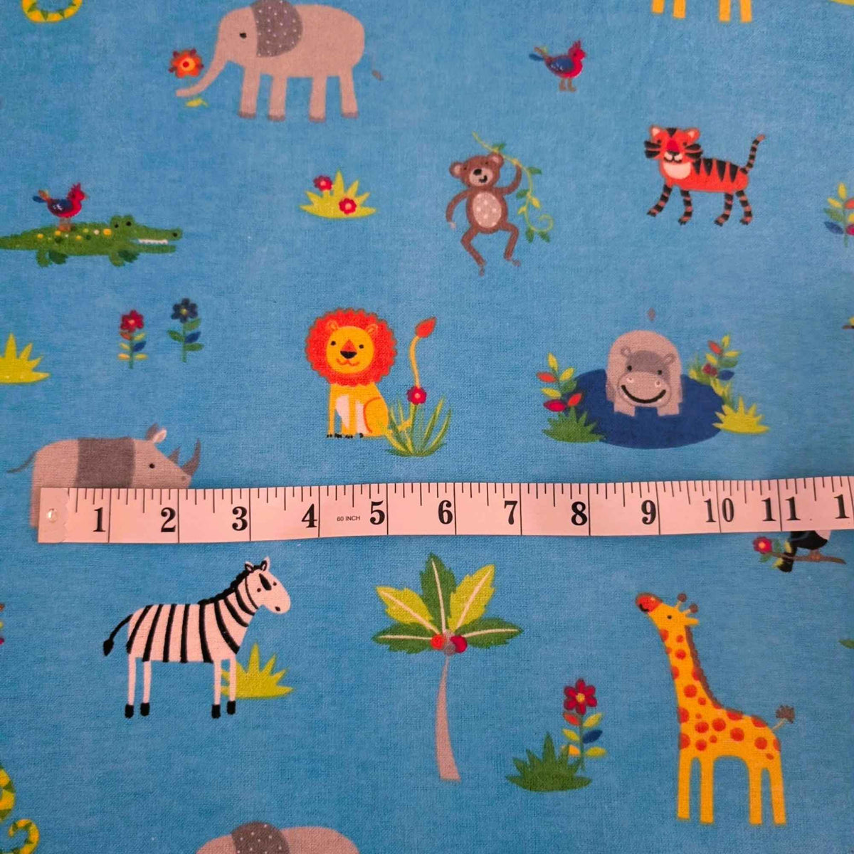 Blue fabric with a colorful safari animal print, shown with a wooden ruler across the middle to indicate scale. The print features cartoon animals including an elephant, lion, zebra, giraffe, tiger, crocodile, monkey, hippo, and palm tree.
