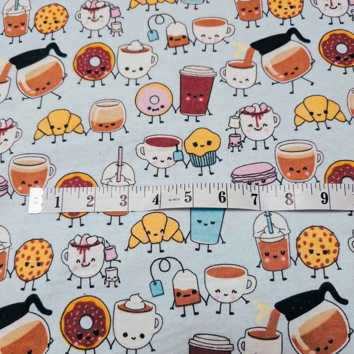 A close-up of a mint green fabric printed with a repeating pattern of cute, anthropomorphic coffee-themed characters, with a white measuring tape ruler placed horizontally across the center to indicate scale.