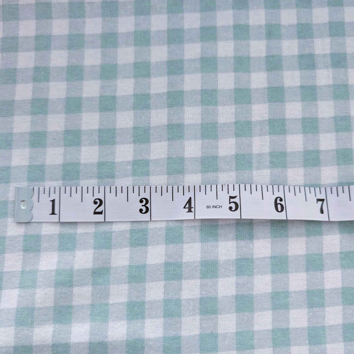 A close-up of a light blue and white gingham fabric with a white measuring tape ruler laid horizontally across the center for scale.